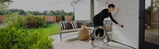 A woman opening the cover of a matte black house hydrant v1+ and carrying a sage green garden hose with a wide grip vb connector on one end and a aquor hose nozzle on the other.
