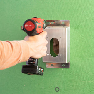 A hand using a power drill to secure the stainless steel mounting box v2+ to sheathing with screws.
