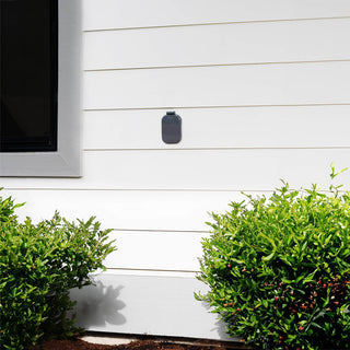 A slate gray house hydrant v2+ with the cover closed on the white lap siding of a home.