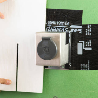 A stainless steel mounting box V1+ with a house hydrant v1+ installed onto sheathing, weatherproofed with zip system flashing. A board is held up to display what siding might look like.