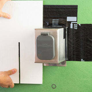 A stainless steel mounting box V2+ with a house hydrant v2+ installed onto sheathing, weatherproofed with zip system flashing. A board is held up to display what siding might look like.