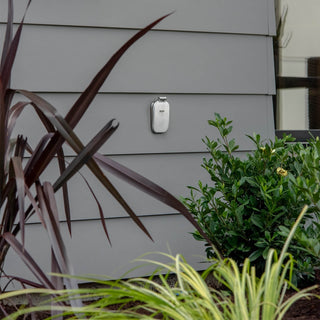 A brushed stainless house hydrant v2+ with the cover closed on the gray lap siding of a home, surrounded by plants.