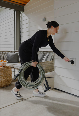 A woman opening the cover of a matte black house hydrant v1+ and carrying a sage green garden hose with a wide grip vb connector on one end and a aquor hose nozzle on the other.