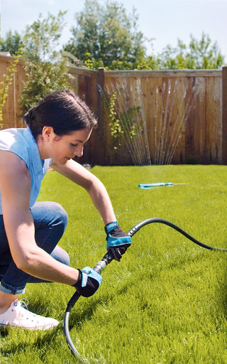 A woman wearing utility gloves connects a black hose adapter to turn on her sprinkler in her backyard.