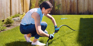 A woman wearing utility gloves connects a black hose adapter to turn on her sprinkler in her backyard.