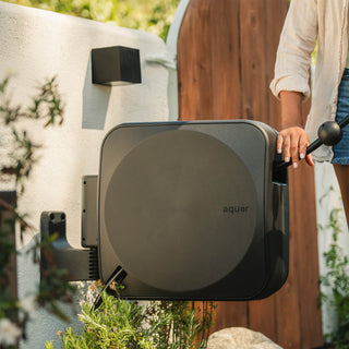 A person pulls a black Aquor Retractable Hose Reel hose in a garden setting installed on a white stucco wall. 