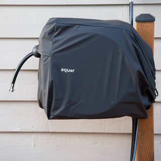 A black retractable hose reel with a branded 'aquor' cover installed on a brown wooden post against a beige sided home.