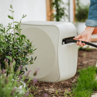 A person is pulling a black hose out from a cream-colored Aquor Retractable Hose Reel, set in a garden environment with green plants and mulch.