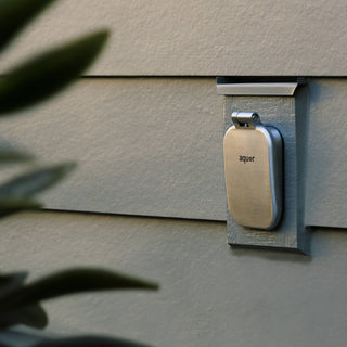 A brushed stainless house hydrant v2+ with the cover closed on the gray lap siding of a home.