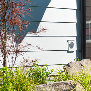 A brushed stainless house hydrant v2+ installed on lap siding with a mounting block, surrounded by plants.