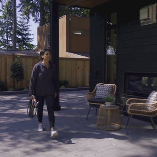 A woman walks across a back porch carrying an Ultralight Garden Hose. She plugs in the Wide Grip VB Hose Connector and hose into the Matte Black House Hydrant V1+ installed on black lap siding.