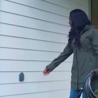 A woman plus in an angled hose connector and hose into a slate gray house hydrant v2+, then water some plants in the yard. 