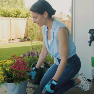 A woman wearing black and blue gardening gloves patting dirt down into a pot filled with yellow, pink, and orange flowers. In the back a Matte Black House Hydrant V1+ with a Comfort Grip VB Hose Connector plugged in. 