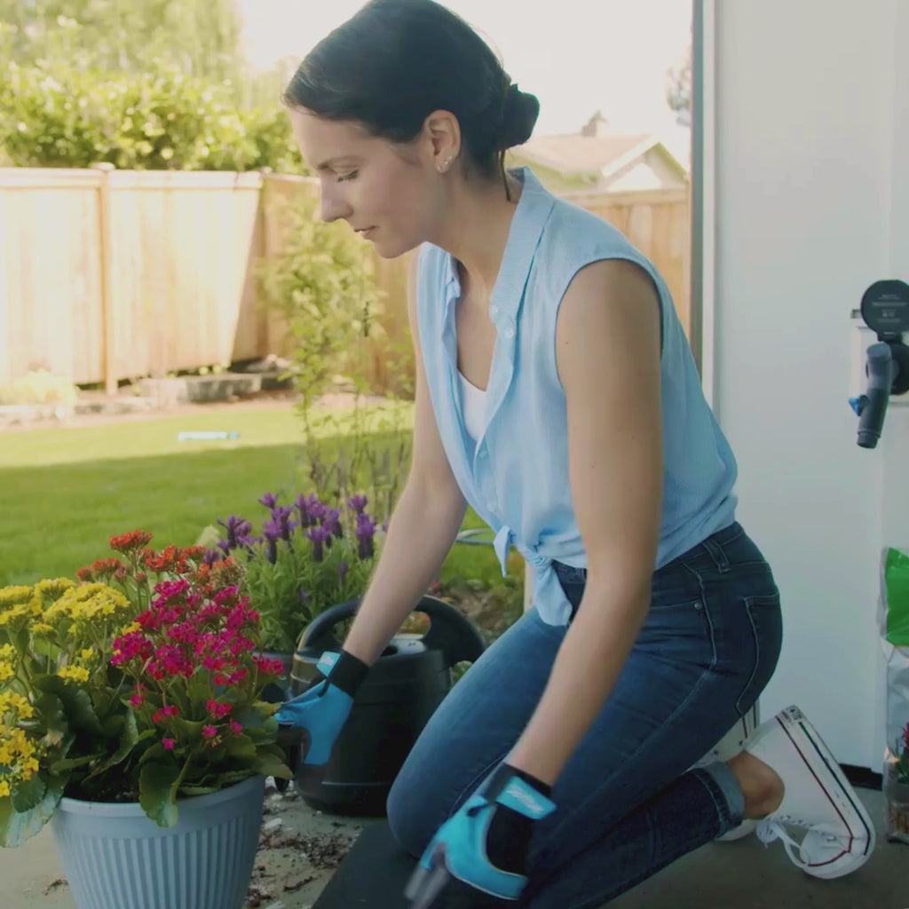 A woman wearing black and blue gardening gloves patting dirt down into a pot filled with yellow, pink, and orange flowers. In the back a Matte Black House Hydrant V1+ with a Comfort Grip VB Hose Connector plugged in. 