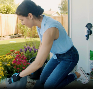 A woman wearing black and blue gardening gloves patting dirt down into a pot filled with yellow, pink, and orange flowers. In the back a Matte Black House Hydrant V1+ with a Comfort Grip VB Hose Connector plugged in.