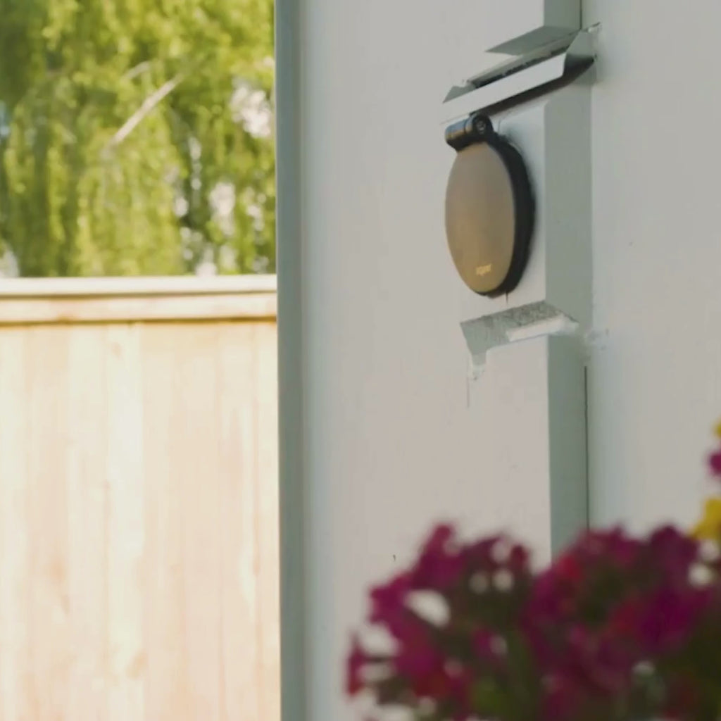 A matte black house hydrant v1+ installed on white lap siding. Closed cover shot, woman plugging in with comfort grip vb hose connector and filling up a watering can then watering a potted plant. 