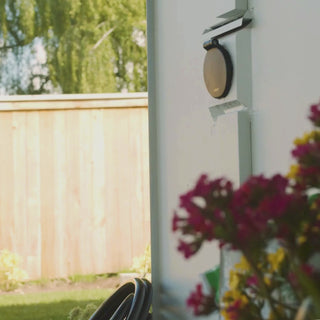 A front view of the Matte Black House Hydrant V1+ installed in white lap siding with the cover closed. Surrounded by pink and purple flowers. 