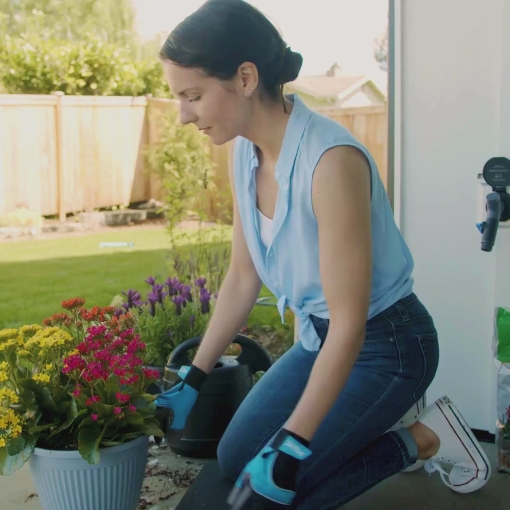 A woman wearing black and blue gardening gloves patting dirt down into a pot filled with yellow, pink, and orange flowers. In the back a Matte Black House Hydrant V1+ with a Comfort Grip VB Hose Connector plugged in.